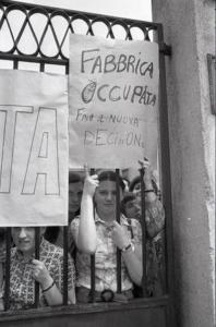 Occupazione della Confezioni Galimberti: primo piano di una delle lavoratrici dietro i cancelli, che mostra un cartello con la scritta "Fabbrica occupata fino a nuova decisione"