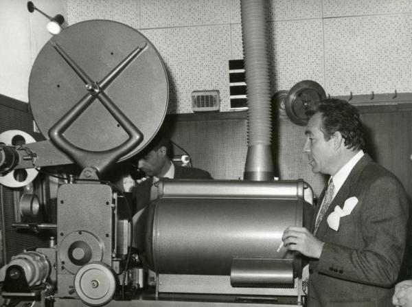 Sul set del film "Il fischio al naso" - Tognazzi, Ugo, 1967 - Ugo Tognazzi, a destra con una sigaretta in mano, rivolge lo sguardo verso una grande macchina da presa. In secondo piano è presente un operatore non identificato.
