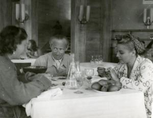 Sul set del film "La famiglia Brambilla in vacanza" - Regia Carl Boese, 1941 - Carl Boese e l'attrice Helen Lüber, a destra, sono seduti a un tavolo imbandito. A sinistra, seduta, una donna non identificata di spalle che si rivolge al regista.
