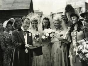 Scena del film "Il fanciullo del West" - Regia Ferroni, Giorgio, 1942 - Erminio Macario, a sinistra, mostra un cappello mentre un'attrice non identificata gli porge un mazzo di fiori. Dietro, tre attrici non identificate vestono i panni di spose.