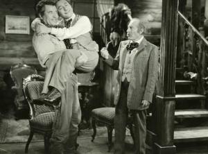 Scena del film "Il fanciullo del West" - Regia Ferroni, Giorgio, 1942 - Un attore non identificato tiene in braccio Erminio Macario che gli stringe le braccia al collo. Intanto, un attore non identificato a destra tiene in mano una pipa e li osserva.