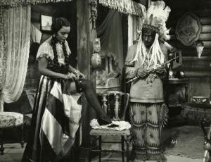 Scena del film "Il fanciullo del West" - Regia Ferroni, Giorgio, 1942 - Un attore non identificato, a destra, tiene in mano un'ascia e guarda Rossana Podestà che, appoggiando il piede destro su una sedia, si sistema la calza e la giarrettiera.
