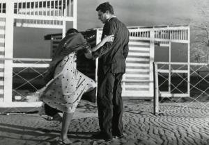 Scena del film "Febbre di vivere" - Regia Gora, Claudio, 1953 - Su una spiaggia, Marcello Mastroianni sostiene con il braccio sinistro un'attrice non identificata intenta a sistemarsi una scarpa.