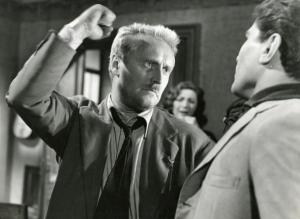 Sul set del film "Il ferroviere" - Germi, Pietro, 1956 - Pietro Germi, a sinistra, solleva il pugno destro in direzione di un attore non identificato, a destra. In secondo piano, un'attrice non identificata si dispera.
