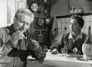 Sul set del film "Il ferroviere" - Germi, Pietro, 1956 - In una cucina, seduti a un tavolo: Pietro Germi è intento ad accendersi un sigaro con dei fiammiferi mentre Luisa Della Noce lo osserva preoccupata.
