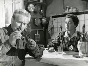 Sul set del film "Il ferroviere" - Germi, Pietro, 1956 - In una cucina, seduti a un tavolo: Pietro Germi è intento ad accendersi un sigaro con dei fiammiferi mentre Luisa Della Noce lo osserva preoccupata.
