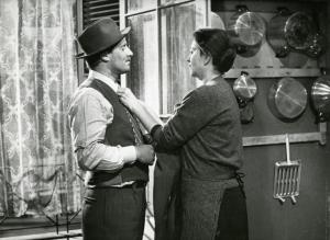 Sul set del film "Il ferroviere" - Germi, Pietro, 1956 - Davanti alla finestra di una cucina: Luisa Della Noce, a destra, sistema la cravatta di Pietro Germi, a destra, che la guarda assorto.

