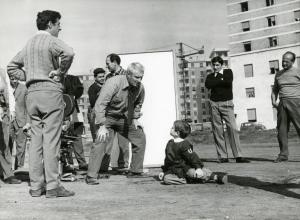 Sul set del film "Il ferroviere" - Germi, Pietro, 1956 - Circondati da attori non identificati, tra cui si vede anche una cinepresa, Pietro Germi, al centro, accovacciandosi si avvicina a Edoardo Nevola, a sinistra, seduto a terra.
