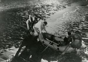 Scena del film "La figlia di Mata Hari" - Merusi, Renzo, 1954 - Un'attrice non identificata è seduta su un gommone mentre, due attori non identificati, a destra, insieme a Erno Crisa, a sinistra, sono intenti a trascinare il mezzo in acqua.
