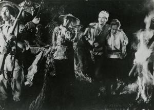 Scena del film "La figlia di Mata Hari" - Merusi, Renzo, 1954 - Sorretto da un attore non identificato, Frank Latimore, con una benda sulla fronte, guarda in direzione di attori non identificati con indosso abiti militari e cappelli asiatici.
