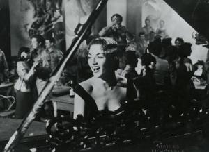 Scena del film "La figlia di Mata Hari" - Merusi, Renzo, 1954 - Primo piano di un 'attrice non identificata che, al centro dietro a un pianoforte con la bocca spalancata, sorride. In secondo piano attori non identificati conversano e danzano.
