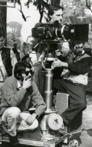 Sul set del film "Francesca è mia" - Russo, Roberto, 1986 - Roberto Russo, a sinistra seduto sulla macchina da presa con una mano sotto il mento, osserva davanti a lui. Dietro e di fianco alla macchina da presa, altri operatori non identificati.